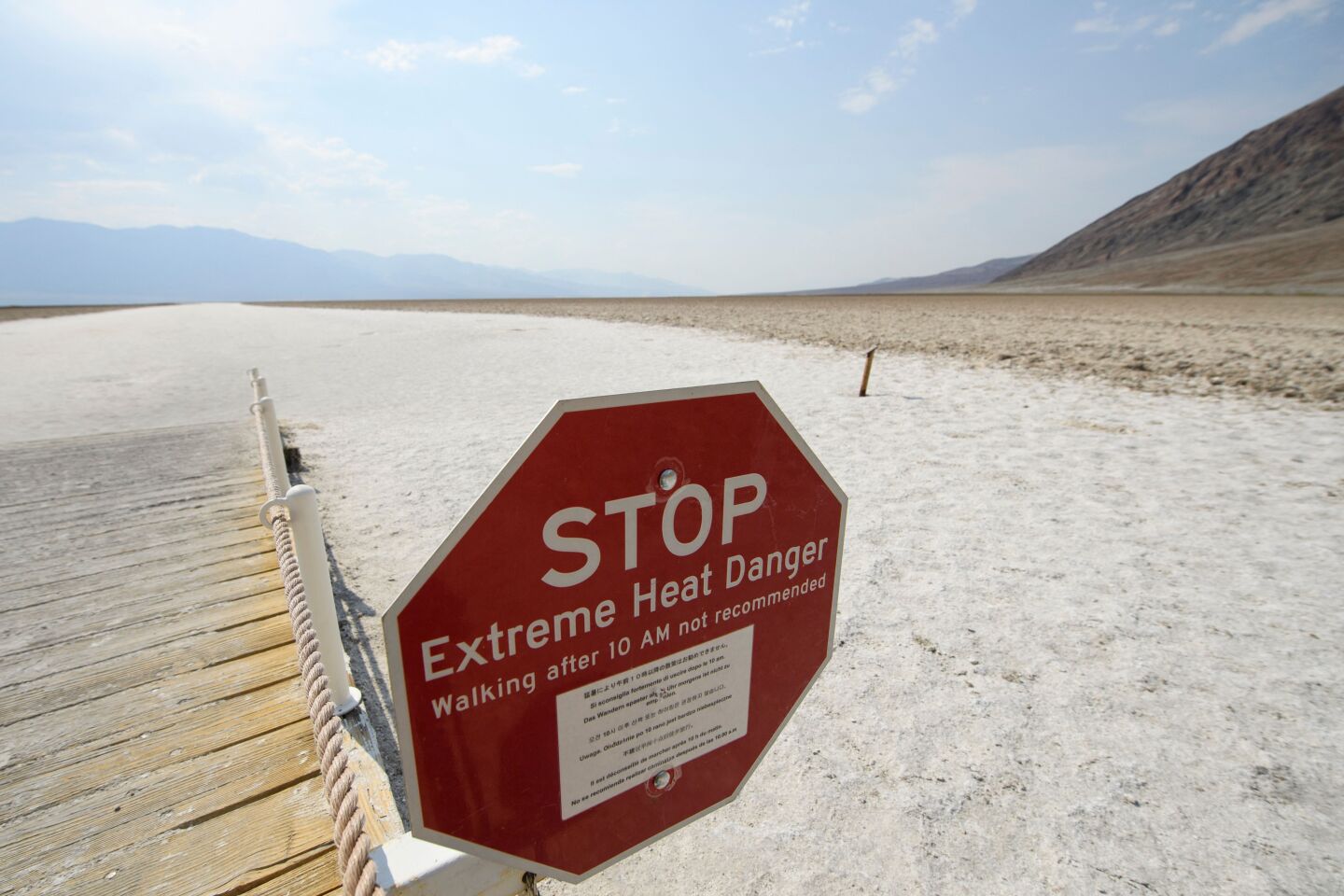 A red stop sign warns: "Extreme Heat Danger. Walking after 10 AM not recommended." It is surrounded by a wood walkway, sand and barren hills.