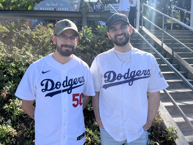 Two men wearing beards and Dodgers jerseys pose outside by a flight of stairs.