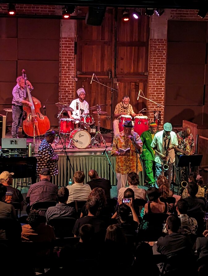 Eight musicians in the Pan-Afrikan Peoples Arkestra perform on and inf ront of a stage as an audience of people watch. A few people seated in the audience are recording on the phones.