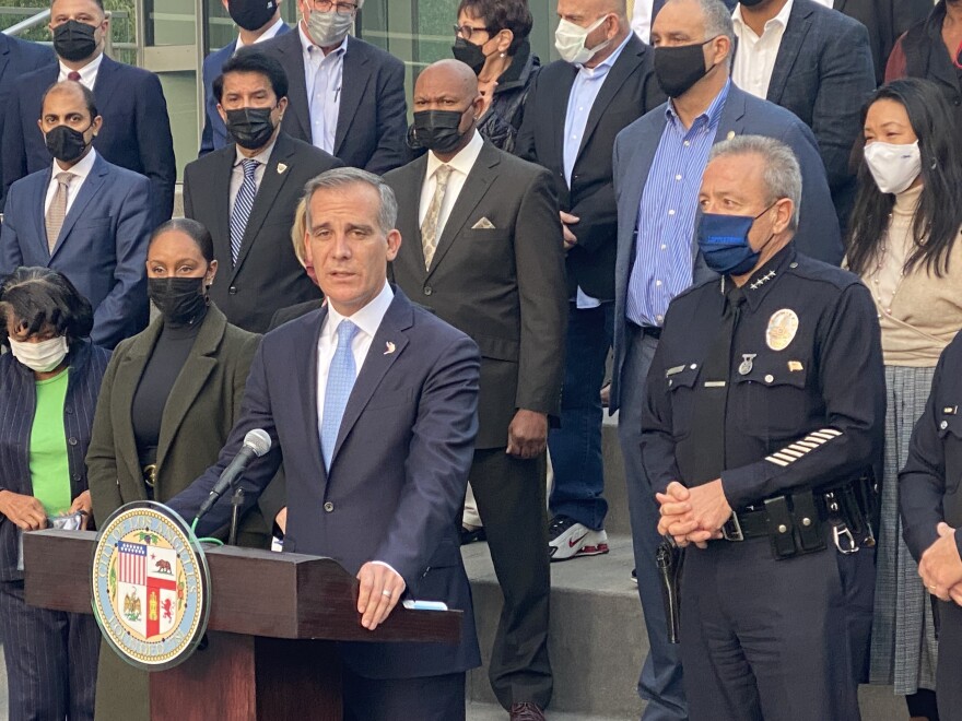 L.A. Mayor Eric Garcetti speaking a podium with the city seal affixed to the front. He is flanked by a masked LAPD Chief Michel Moore, and more than a dozen other law enforcement and business leaders are behind him.