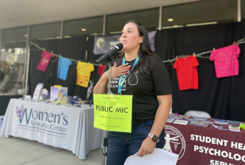A woman with long dark hair stands behind a microphone labeled "public mic." She holds a white sheet of paper in her left hand. Her right hand is placed over heart.