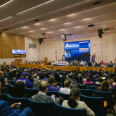 A large auditorium where a large crowd sits in blue chairs facing a dais with a large screen above. 