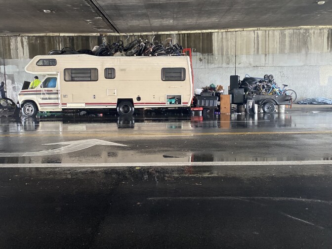 An RV is park under a freeway overpass with bicycles piled on top of it and on a trailer behind it.