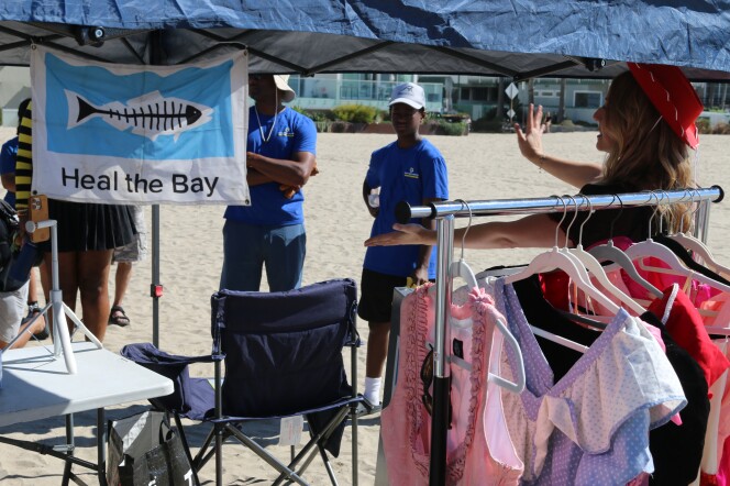 A view of people standing under a pop-up canopy with a Heal the Bay sign and a rack of clothing in view. To the right is Lexy Silverstein, a white woman with a red hat on, as she talks to people watching.