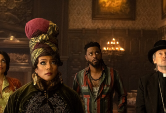 Rosario Dawson, Tiffany Haddish, LaKeith Stanfield, and Owen Wilson stand in an ornate living room with carved wooden wall panels with candelabras hanging off those walls, washing the room in a dim golden glow with a slight haze of dust. They all stare up at something on the ceiling or high on the wall in front of them. Stanfield and Wilson look on seemingly worried while Dawson and Haddish look more unphased by what they are witnessing. Dawson wears a an orange sweater over a green collared shirt. Haddish wears a tall headscarf and heavy dark green cloak. Stanfield wears a green, red, and blue striped button down. Wilson wears a black fedora-like hat and priest suit. 