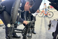 Police officers examine a folded electric bike's gears in a parking lot as another writes on a clipboard.
