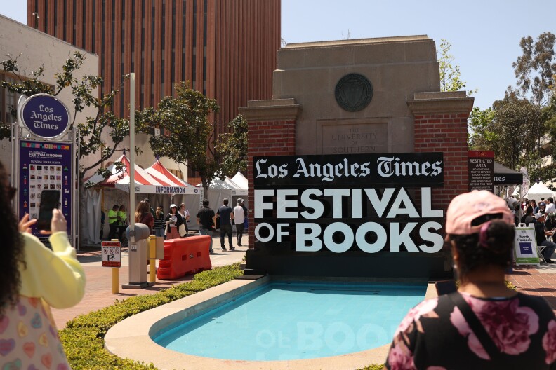 A giant black and white sign reading "Los Angeles Times Festival of Books" is displayed over a stone and brick reflecting pool. Two people can be seen taking photos of it with their phones.