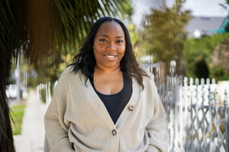 A portrait of a Black person standing outside in front of a palm tree and white fence. They are wearing a tan cardigan while smiling at the camera.