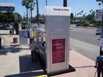 A person stands in the shade provided by a bus shelter as other people stand and walk around at a transit station next to a street.