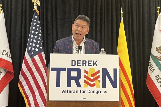 A Vietnamese American man in his 40s wearing a blue suit checked with red answers stands at a podium with a sign that reads "Derek Tran: Veteran for Congress."