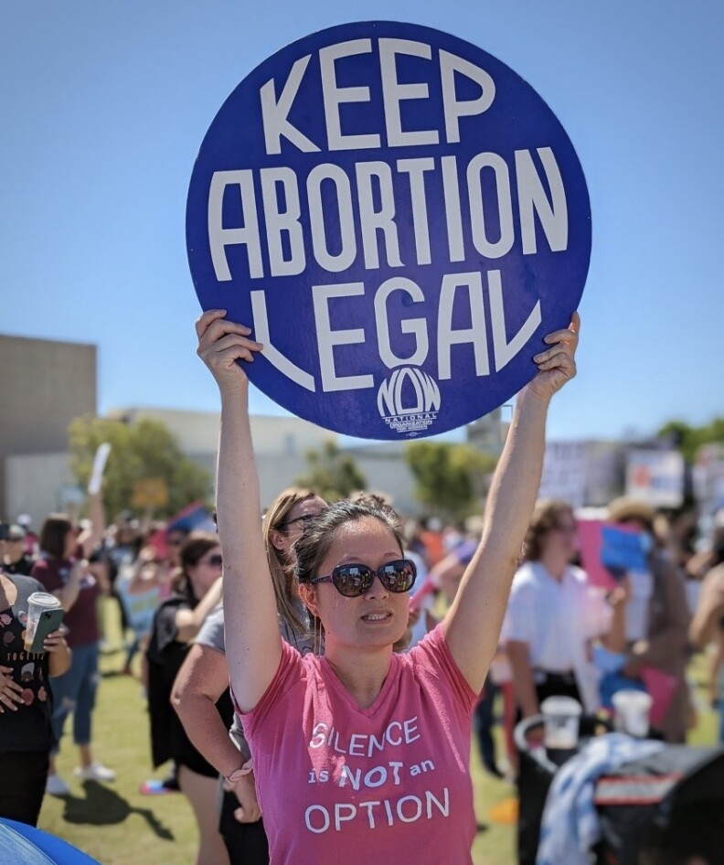An Asian American woman wearing sunglasses and a pink shirt that says "Silence Is Not An Option" holds up a sign that reads "Keep Abortion Legal."
