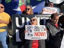 A woman with a medium-light skin tone and dark sun glasses holds a white sign that reads "Overpaid CEO Tax Now! CEOTAX.LA." Behind her, others hold a Unite Here banner. 