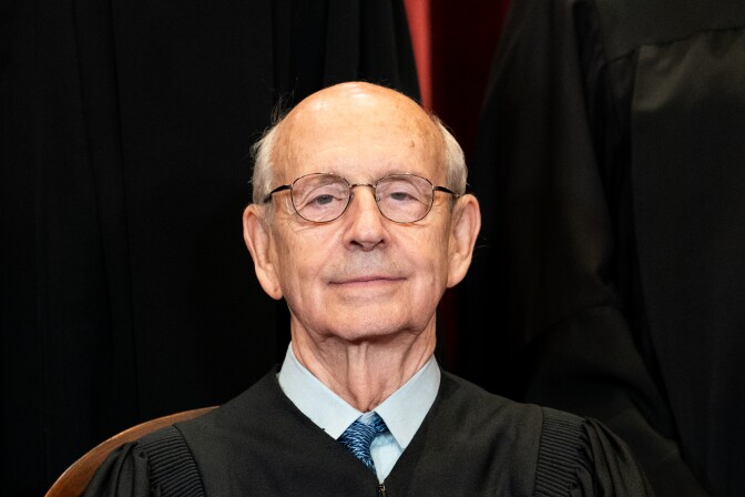 Justice Stephen Breyer, an older white man with grey, thinning hair and wire rim glasses, is shown in a headshot. He wears a blue shirt and tie under his judicial robe. 