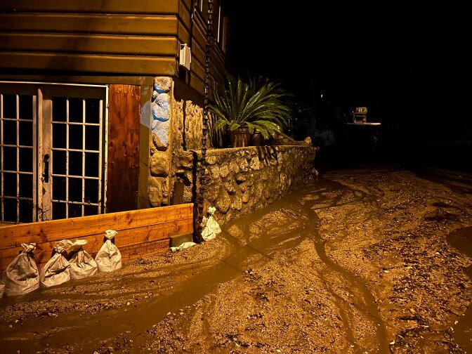 Mud and water flow down a street at night. Sandbags can be seen lining a wooden fence of a home in the background.