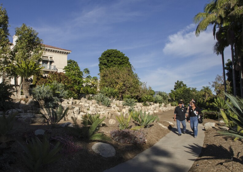 A small group of people walk down a concrete path with large ferns, and cacti on either side with decorative stones to their right. They are approaching a building on the grounds with a balcony and spanish style roof tiles.