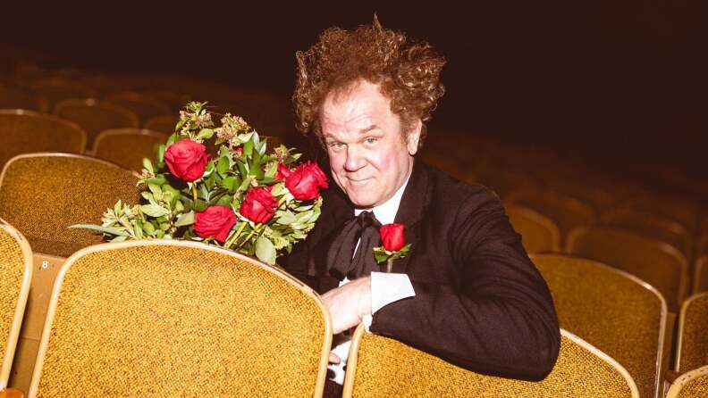 John C. Reilly, wearing a suit, holds roses and smirks at the camera as he sits in yellow theater seats. He's in character as Mister Romantic.