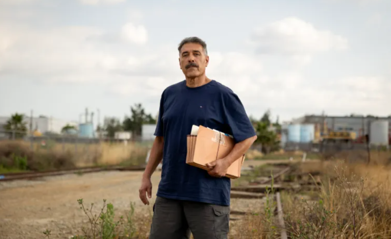 A man with medium skin tone poses for a photo, in front of the nearby plant. He is wearing a dark blue t-shirt and is holding a paper file box.