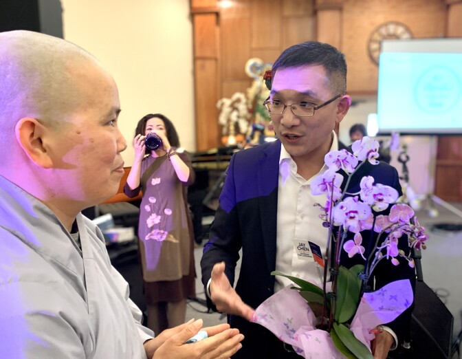 An Asian American man in his 40s wearing glasses and a suit (l.) presents a potted plan to a nun (r.) who wears a smile and a shaven head.
