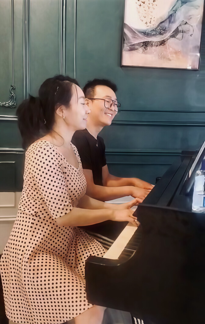 A light-skinned woman with dark hair pulled back wears a pink polka-dot dress. She plays a black piano alongside a light-skinned man with short black hair and glasses, wearing a black T-shirt. Both are smiling in front of a dark teal paneled wall.