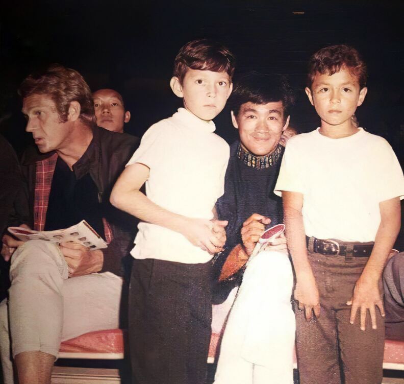 A white blonde man sits on the left a chair. To the right of the image, a smiling Asian man poses with two young fans.
