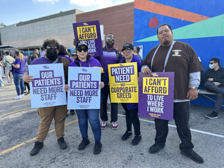 Five people stand holding signs that read "our patients need more staff" "patient need not corporate greed" and "i can't afford to live wehre I work." There are two women with light brown skin, a man with medium brown skin, and man with dark brown skin, and a younger man with light brown skin with curly hair wearing a mask.