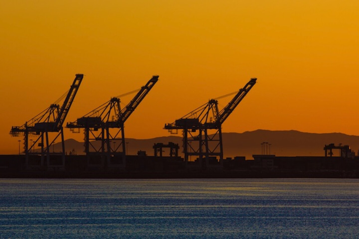 Three large machines are shown on the horizon of the Port of Long Beach. A vibrant orange sunset is in the background, and the blue water is in the foreground.