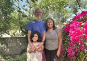 An older male and female couple with brown skin stands with a young boy with long black hair, all smiling for the camera. The man wears a blue t-shirt and the woman a light grey t-shirt. The boy wears an offwhite t-shirt. They are surrounded by trees and bougainvillea. 