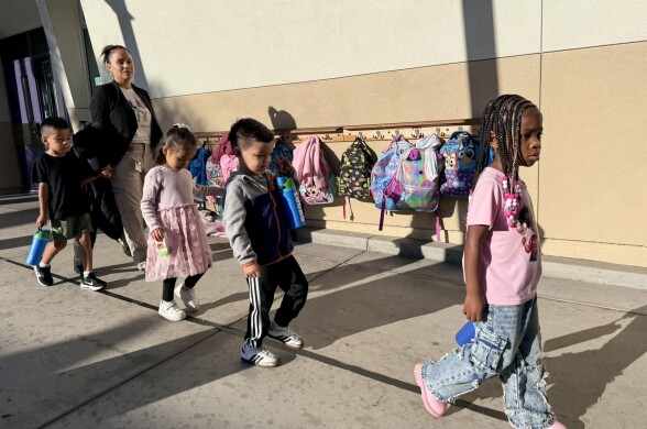 Students with a variety of skin tones walk in a straight line in front of a row of backpacks. 