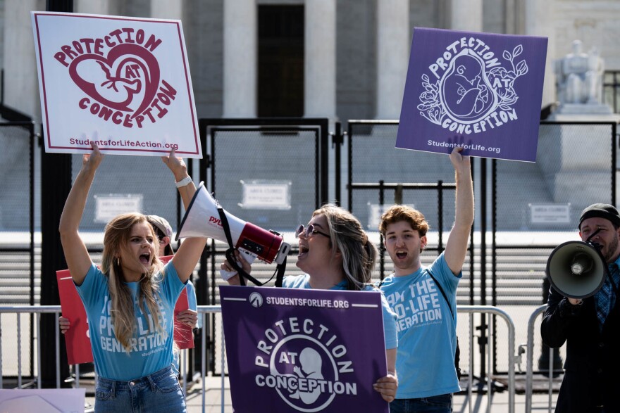Three young white people protest abortion rights outside the U.S. Supreme Court, holding signs that read "protection at conception." One stands behind a podium and speaks into a bullhorn.