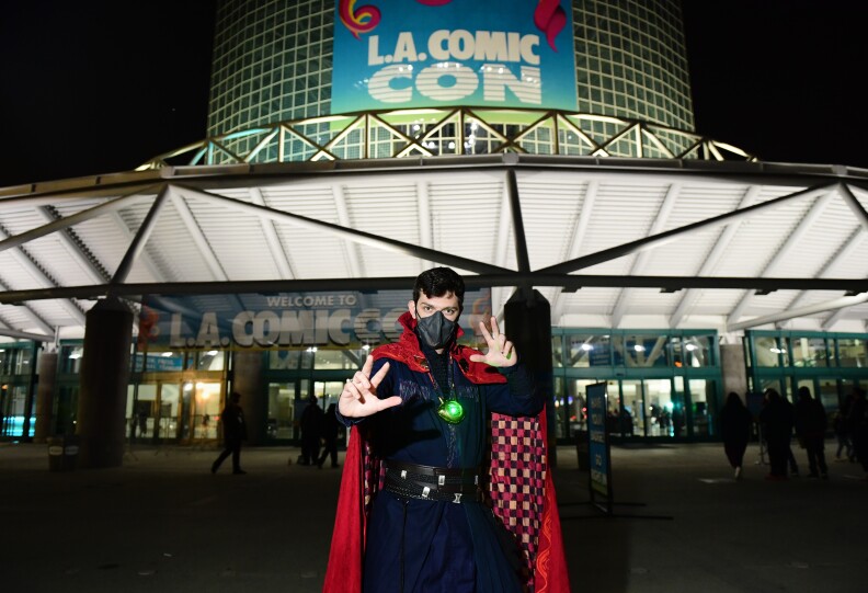 A fan dressed as the film version of Marvel superhero Doctor Strange poses, making magic gestures with his hands, and wears a mask, outside of the L.A. Convention Center. It's night, but the center is illuminated behind him, with large banners reading "Welcome to L.A. Comic Con." A number of passers-by are behind him.