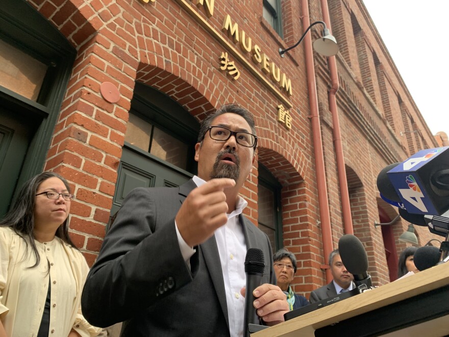 A Korean American man standing at a podium outside a brick building holds up his hand while talking and holding a mic in the other. On the left side of the photo, standing behind him, is a Chinese American woman in a yellow shirt. On the right side of the photo, standing behind him, are a Korean American woman and Chinese American man.