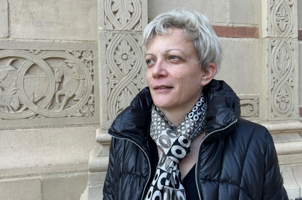 A light-skinned woman with short hair smiles slightly as she gazes in front of her. She is standing in front of an ornate brick, wearing a black jacket and scarf. 