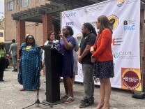 Six people, five women and one man, are lined up behind a black podium with microphones attached. The woman in the center is speaking directly into the microphone with her left hand raised, her finger pointing out towards the unseen audience. They're standing in front of a white banner with several different organization's logos on it.