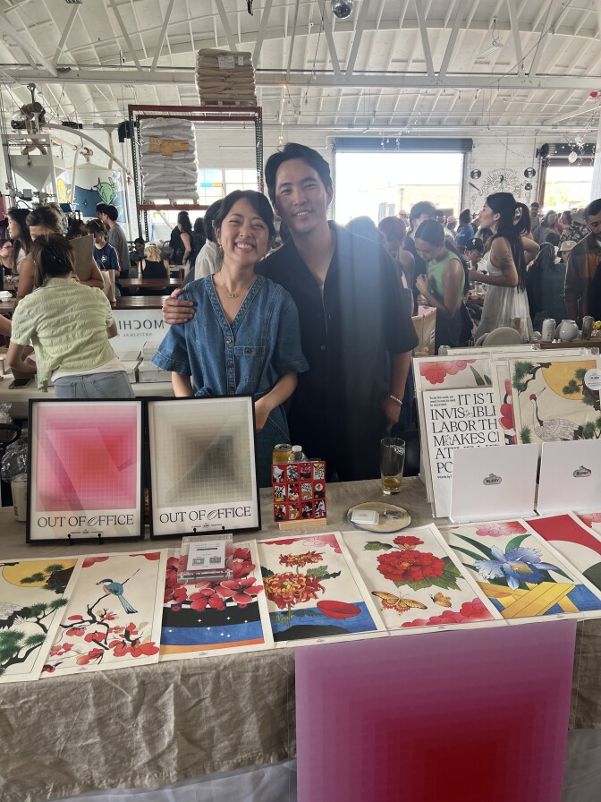 A young Asian man and woman standing behind a table with a lot of art prints.