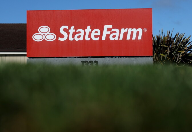 A red State Farm sign stands on a pole behind a blurred lawn, with a house and plant visible in the background.