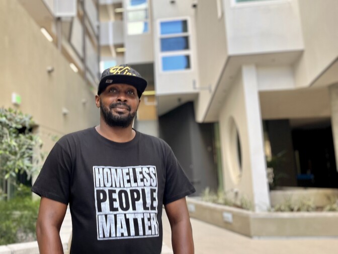A Black man wearing a black "Homeless People Matter" shirt stands in the courtyard of a multi-story residential building.