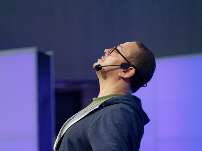 A man wearing glasses, a headset microphone, and a blue zip-up hoodie stands on a stage with his head tilted back and eyes closed. His arms are extended downward and slightly out with palms open in an expressive or mindful gesture.