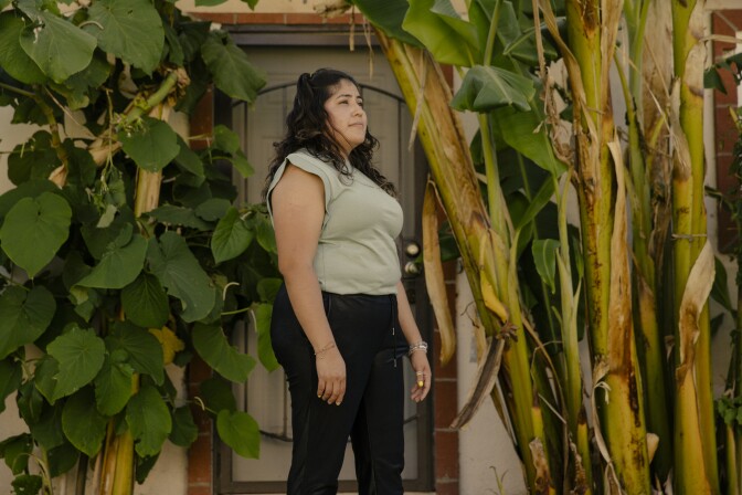A woman with long dark hair wearing a pale green t-shirt and black pants stands in front of a doorway framed by banana plants on one side and a large-leafed plant on the other. She is looking off into the distance. 