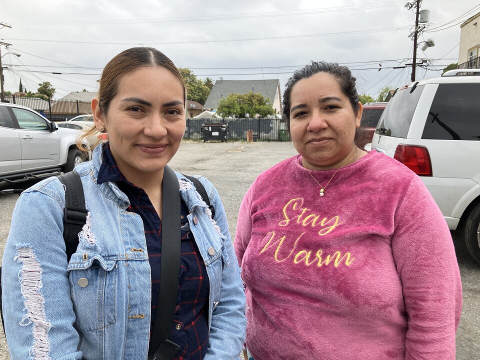 Two female-presenting people, one at left in a blue denim jacket and ponytail, the other at right in a pink sweatshirt that reads "stay warm," with hair pulled back, stand in a parking lot with cars in the background.