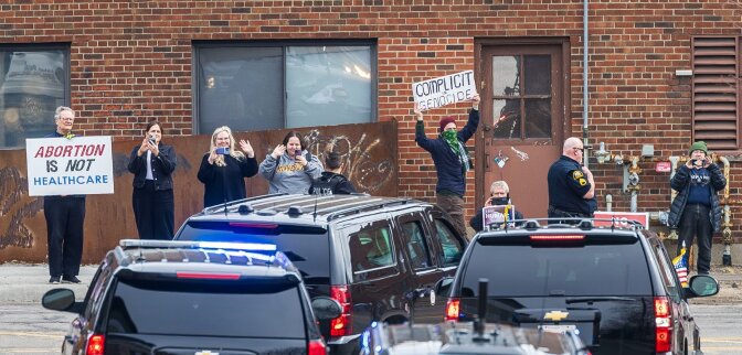 A small group of people, all with light-tone skin, stands along a red brick building as a motorcade of dark cars turns. A few people are waving and taking photos. One person holds a sign reading: "ABORTION IS NOT HEALTHCARE" and another holds a sign overhead that reads: "COMPLICIT in GENOCIDE."