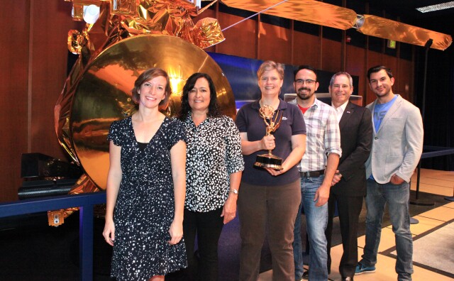 Six people with light skin tone gather for a photo. The third person from the left is holding a trophy.