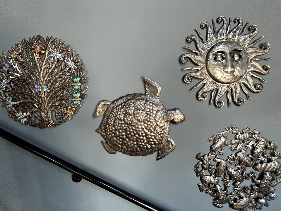 A shot of cut metal wall art hanging. There are four pieces, the one on the left is a tree in a round shape, the middle is a sea turtle, the top right a sun, and the bottom right frogs. 
