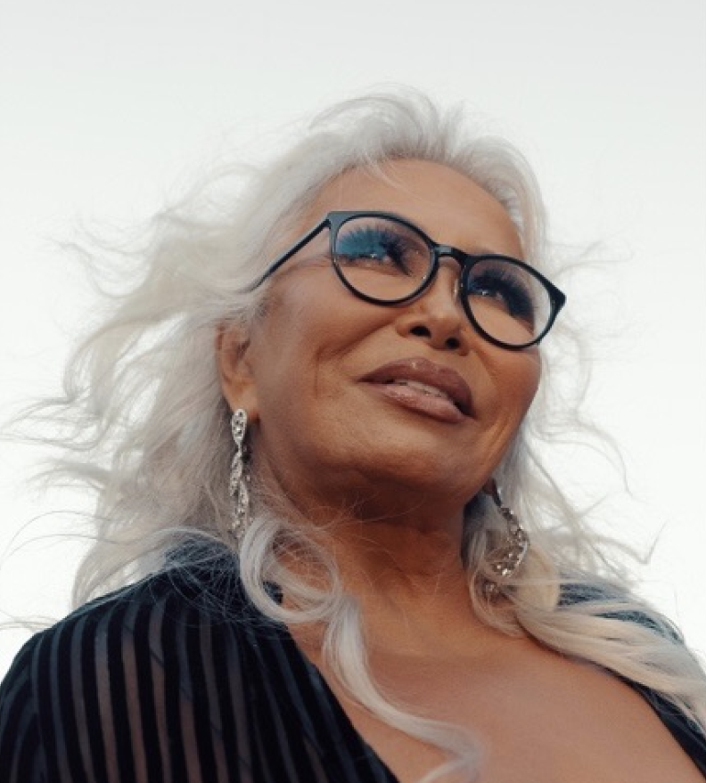 A female-presenting person with medium-brown skin, long white hair, and glasses, wearing a black jacket, looks upward.