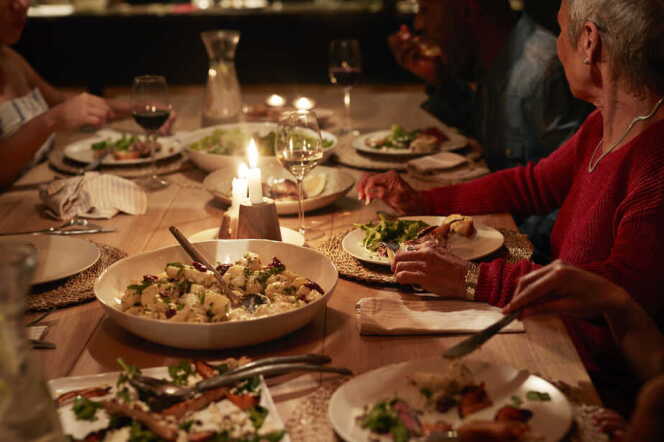 A wood table has food served family style including potatoes and salad with glasses of red and white wine. People sitting at the table have medium-skin tones.