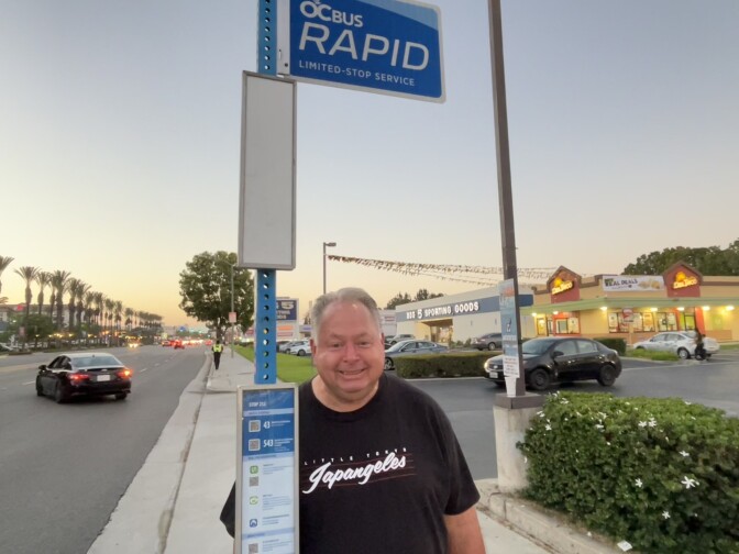 A man with grey hair and fair skin stands in front of a bus stop in Anaheim.