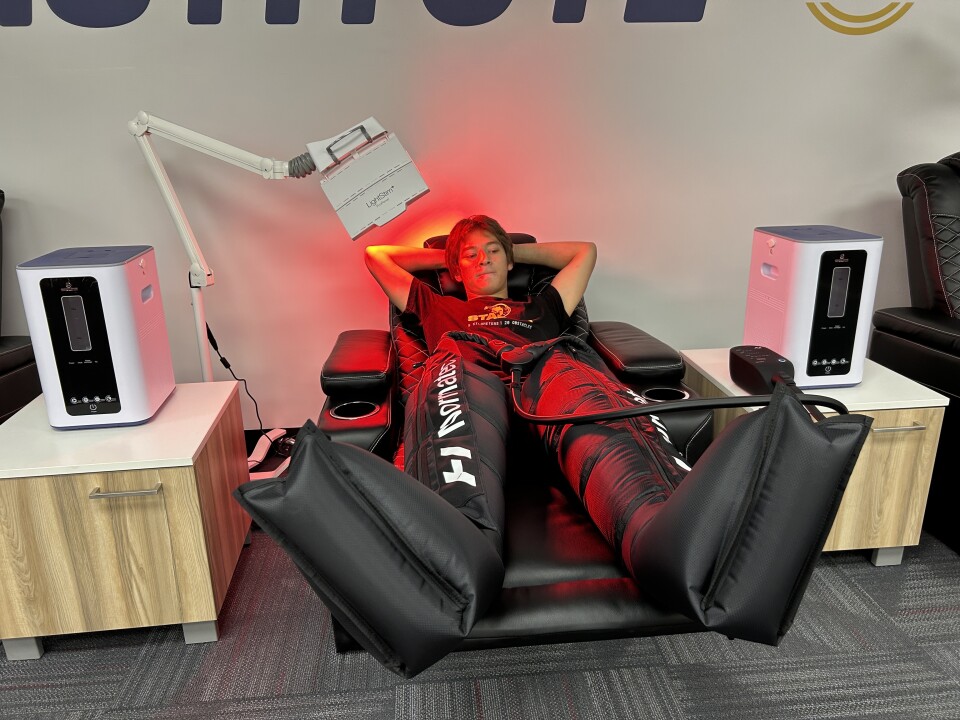 A teenager lies in a black reclining chair with a red light therapy light shining on him and black puffy compression boots on his legs.