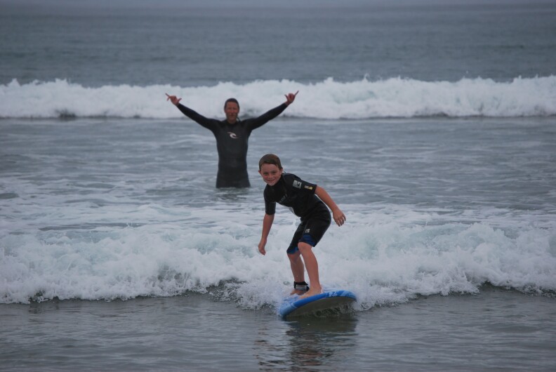 A young boy smiles while surfing a small wave on a blue board. In the background, a tall man with a mustache stands in the water, arms in the air, and flashes hang loose signs with both hands.