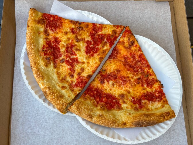 Two classic cheese pizza slices with browned, bubbly crust and patches of tomato sauce, placed on a white paper plate inside a cardboard pizza box.
