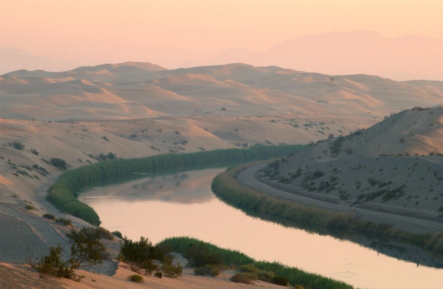 A river curves around between dusty hills. There is thick greenery along the banks. The sky has a yellow-reddish hue.
