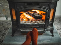 A close-up image of feet in warm-looking red socks in front of a cozy-looking fireplace with a fire roaring. The fireplace looks old and is made of ornate iron. 